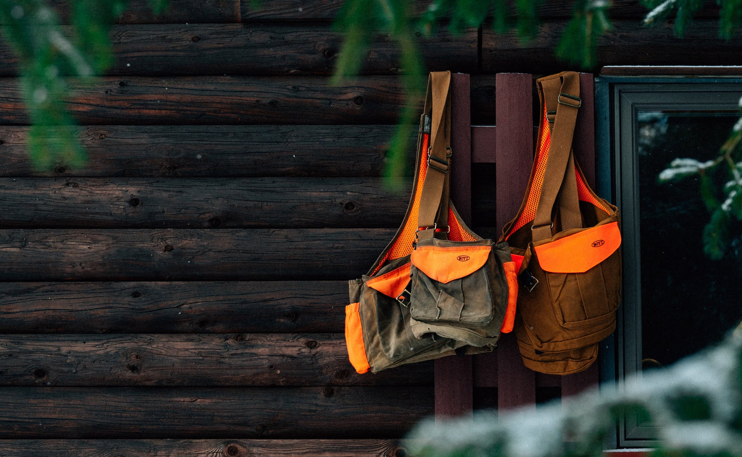 Two Boyt upland hunting vests hanging on a wooden window shutter with snow-covered steps and trees in the foreground.